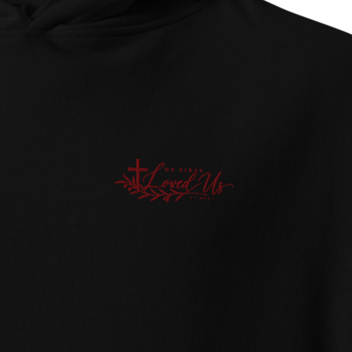 Close-up of black He First Loved Us premium embroidered Christian hoodie with red cross and script chest embroidery.